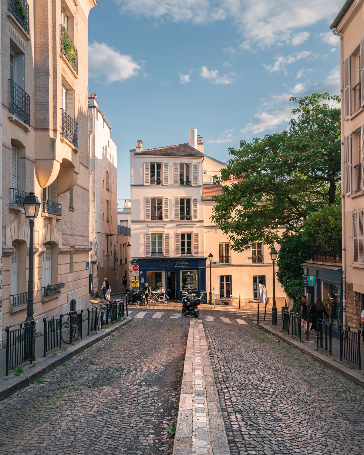 A charming street scene in Paris, featuring cobblestone pavement and elegant buildings under a clear blue sky. The atmosphere is lively, with people and bicycles adding to the urban vibe.