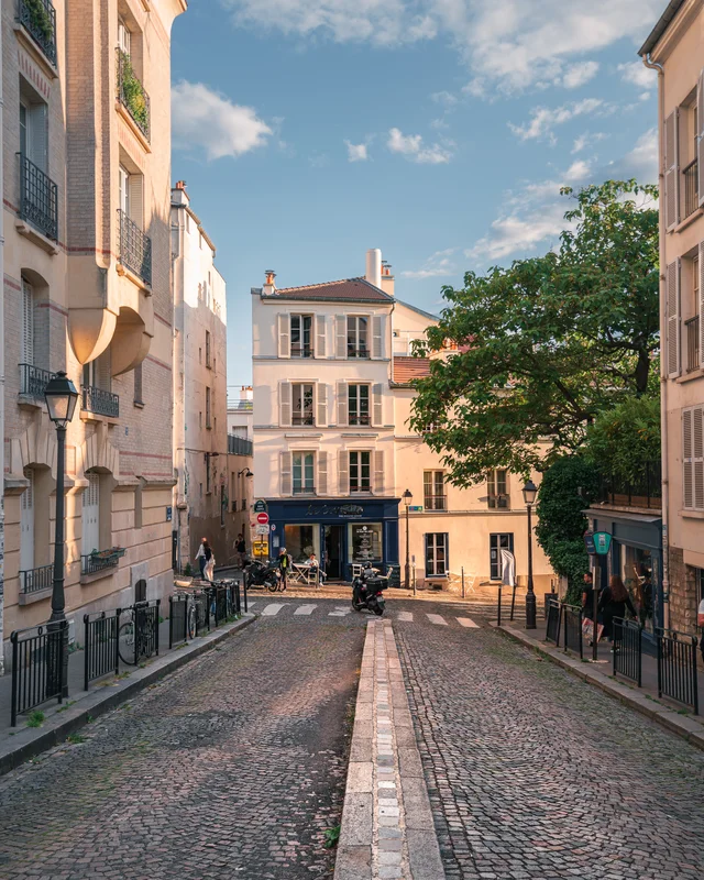 A charming street scene in Paris, featuring cobblestone pavement and elegant buildings under a clear blue sky. The atmosphere is lively, with people and bicycles adding to the urban vibe.