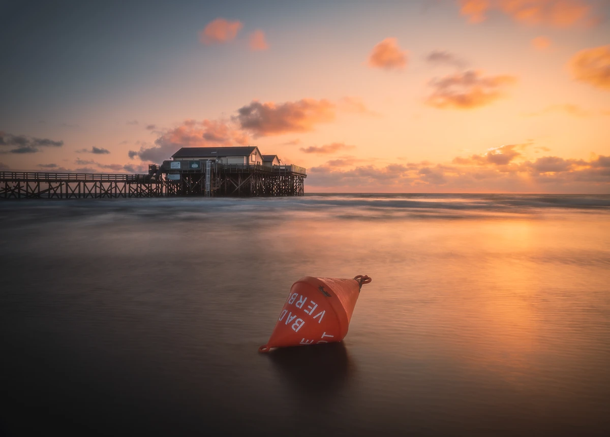 A vibrant sunset casts warm hues over a tranquil ocean, with a buoy resting on the shore in the foreground. The wooden pier stands majestically in the background, creating a serene coastal atmosphere.